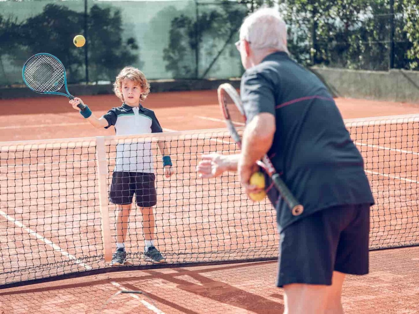 An image of a senior male training in sports clothes explaining how to hit the ball with a racket to the attentive little boy during tennis on an outdoor court.