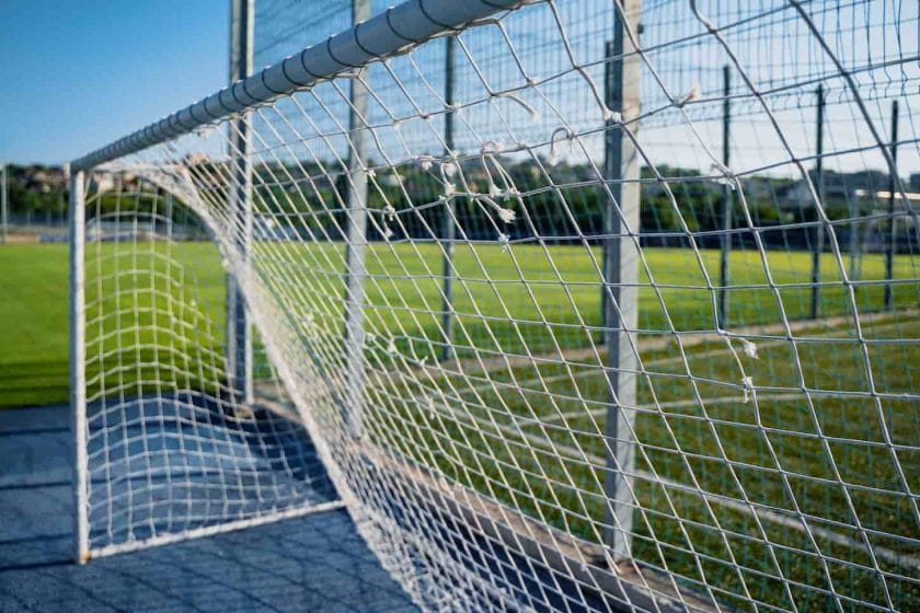 An image of a damaged soccer gate goalpost white rope net with knots and a green field and blue sky in the background.