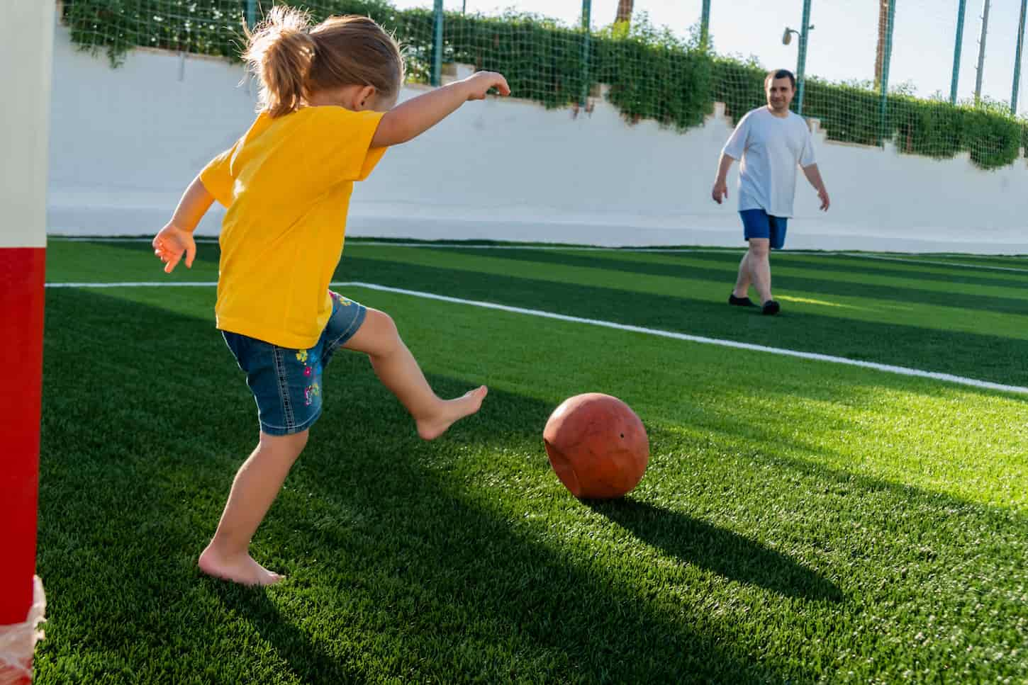 An image of a Cute little girl kicking the soccer ball to her father on the green sports field at sunset time. Kid playing football with her family.