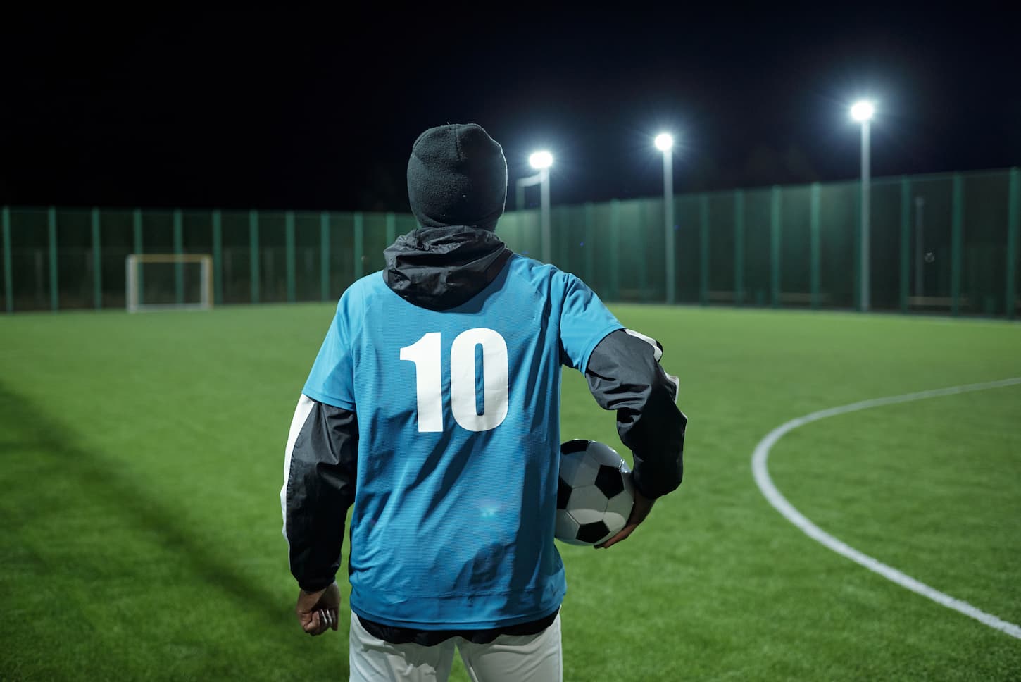 An image of a contemporary professional soccer player in long sleeve uniform and gloves with a ball standing in front of a large football field.