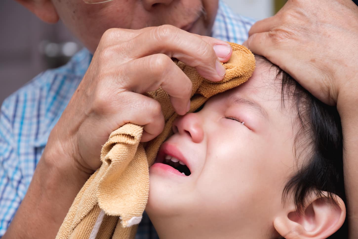 An image of an Asian boy crying out of pain. The child bumps the head against a solid. The baby is bulging. An adult is wrapping an ice cube wrapped in a cold compress on the little boy. Close-up, crying son's face from an injury caused by a home accident. Grandfather takes care of a 2-3-year-old grandson.