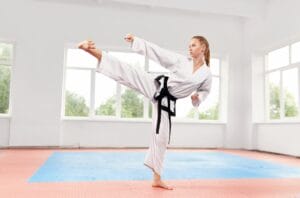 An image of a Sporty strong woman in white kimono with black belt against big window performing martial arts high kick at fight class.